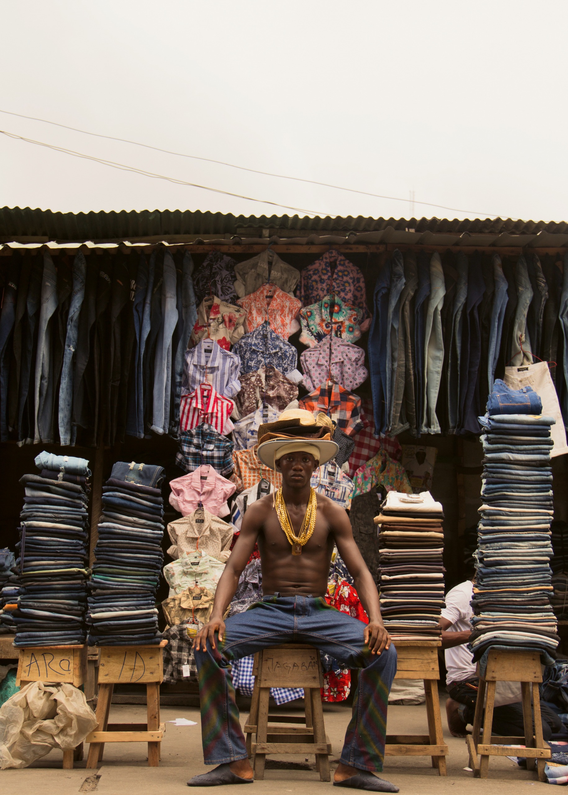 A shirtless man wearing layered gold chains sits on a stool in front of a market stall stacked high with folded jeans and hanging shirts.