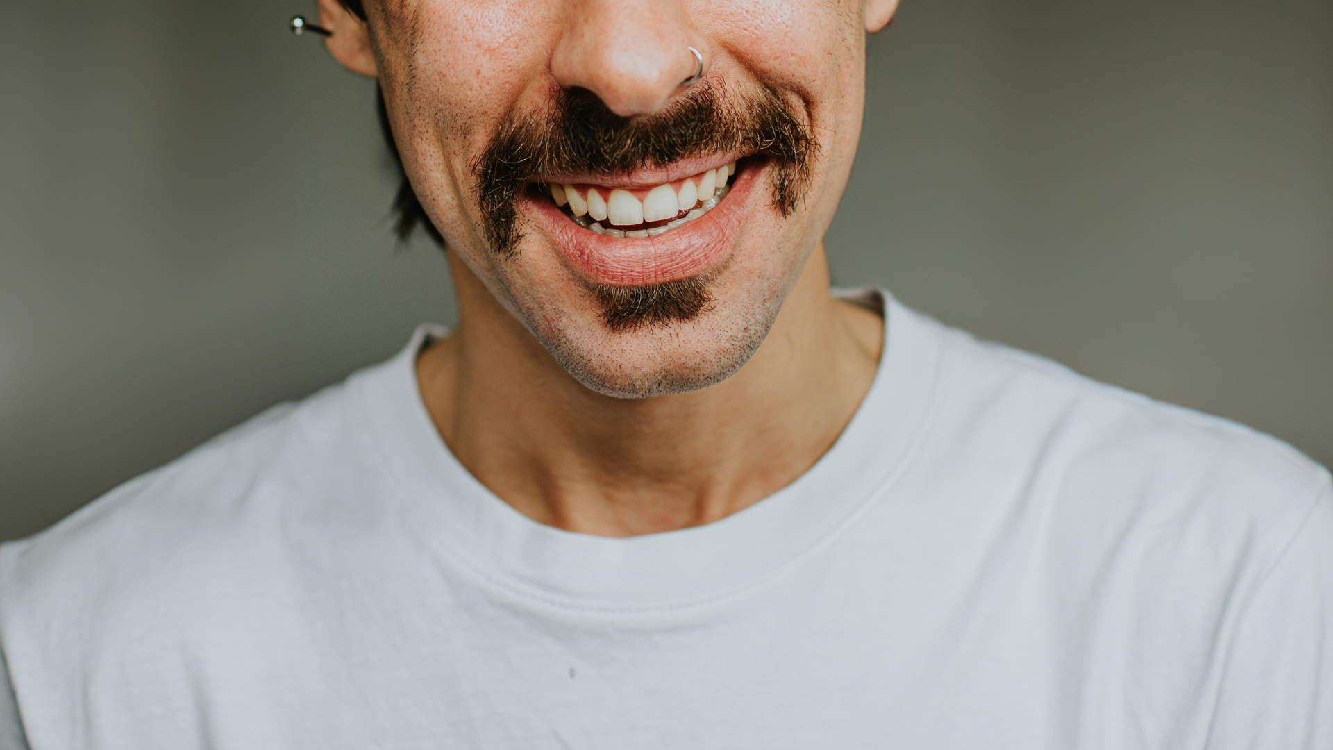 An image of the bottom half of a man's face, showing a dark moustache over a white-toothed smile. The man has an earring bar in his left ear and wears a white shirt