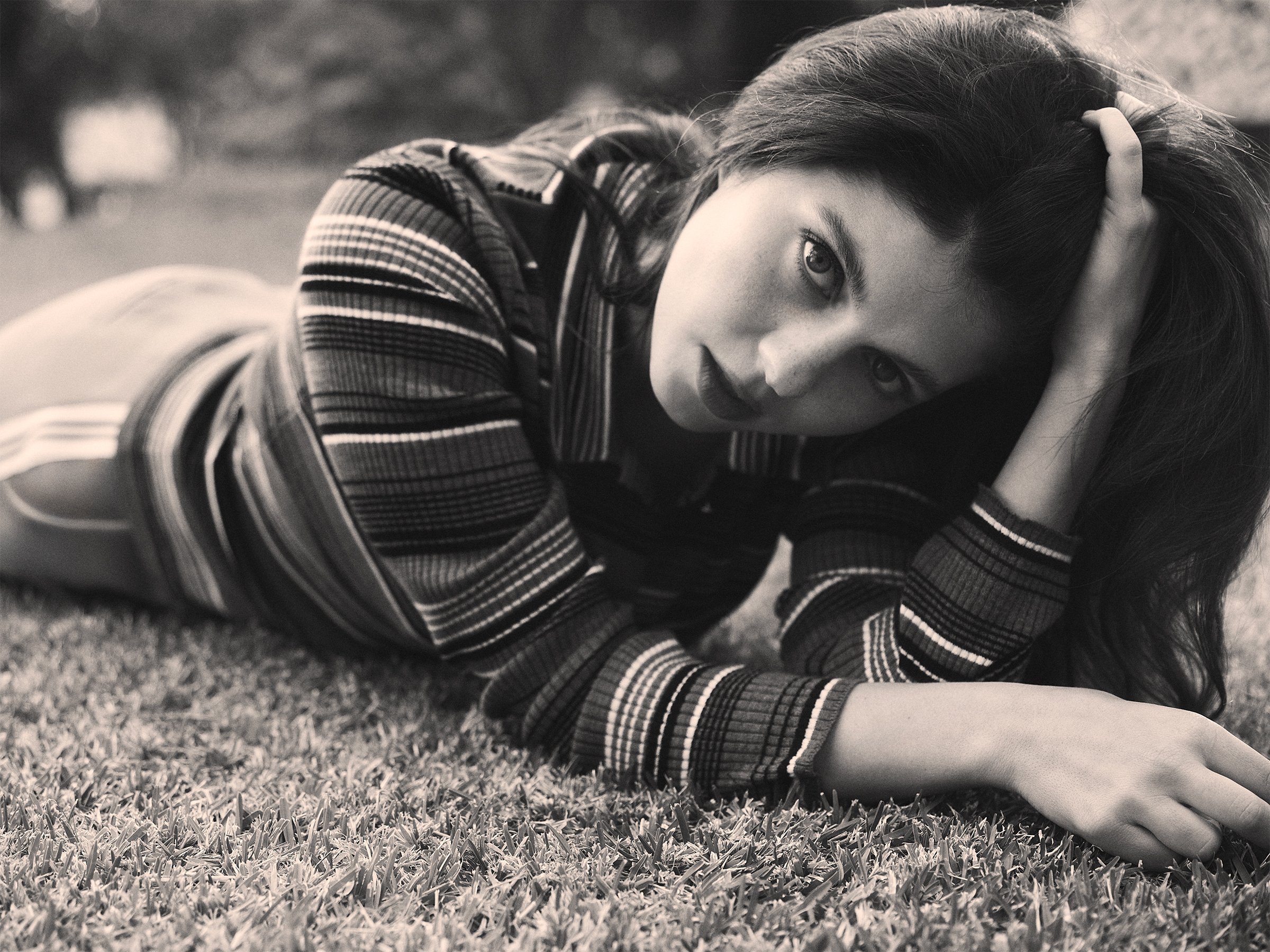Emily Bader is photographed for Who What Wear's In Focus feature lying on the grass. She is running her hand through her hair and wearing a striped cardigan.