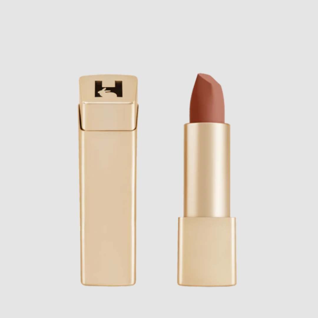 A product shot of the Hourglass Unlocked Soft Matte Lipstick in shade Poppy (a warm, beige brown colour), on a grey background.