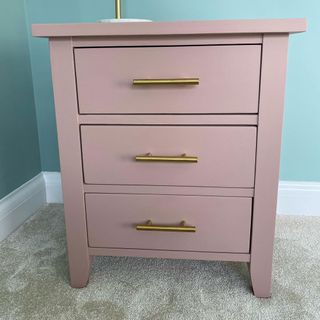 Pink painted side table with brass handles on grey carpet next to blue wall