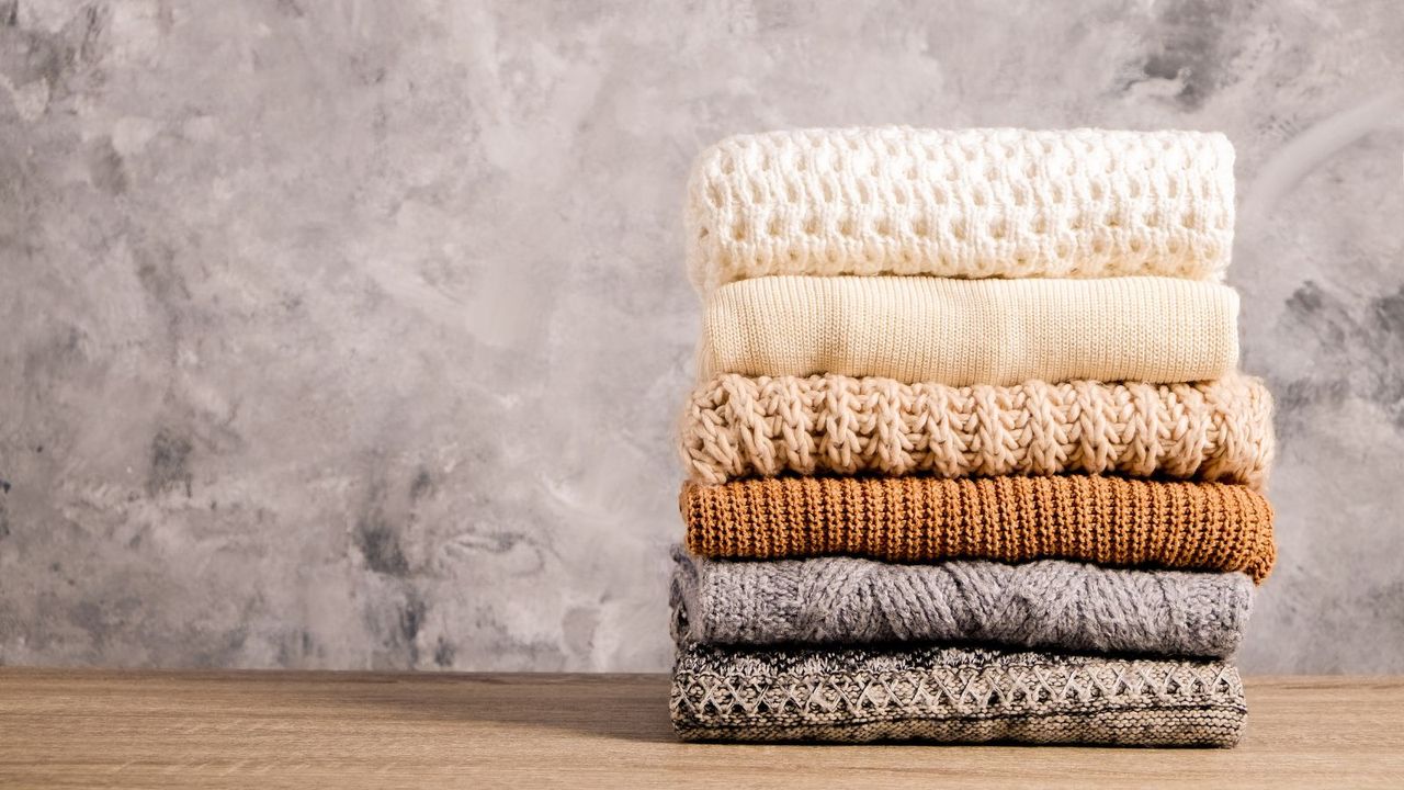 A pile of six sweathers all neatly file folded on a wooden table with a grey backdrop