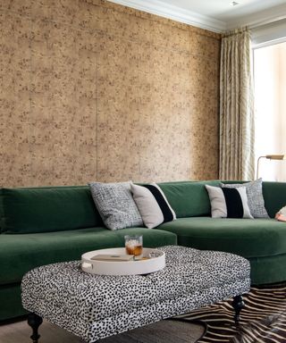 A living area with brown burl wallpaper walls, a deep green curved velvet couch with black and white patterned throw pillows, and a rectangular black and white polka dot ottoman with a circular tray with a glass