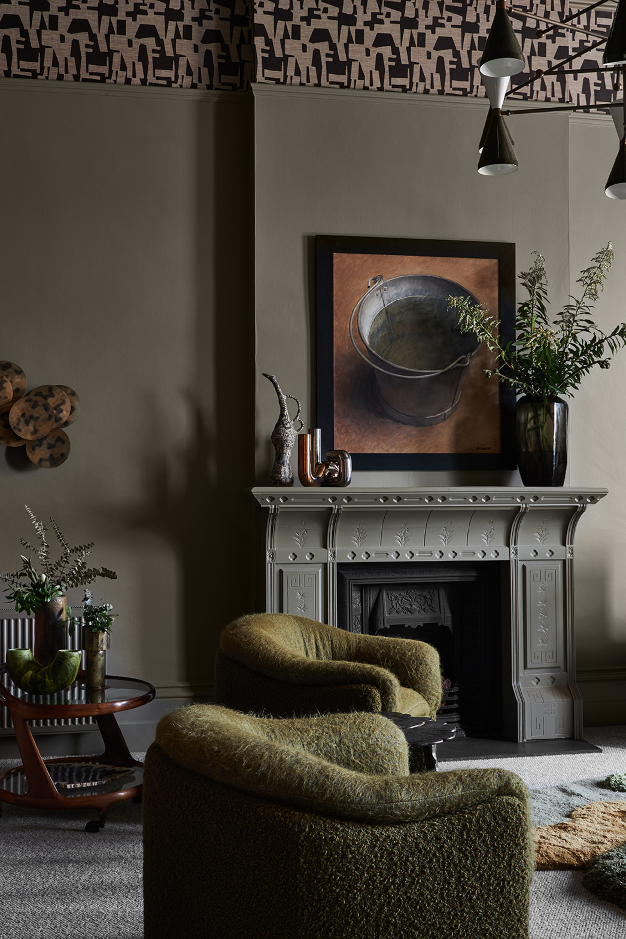 modern living room with dark gray walls, patterned ceiling, green boucle armchairs, an ornate fireplace with artwork and vases styled on the mantel and a small bar cart next to it