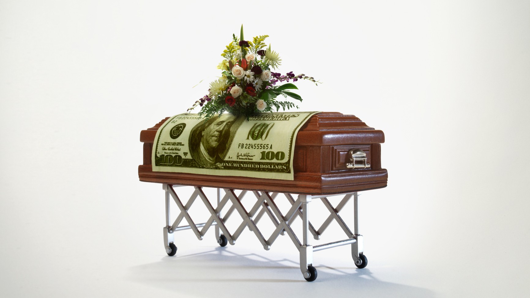 A casket on a guerney is draped in a $100 bill and topped with flowers. Conveys the notion of funerals and the cost of dying.