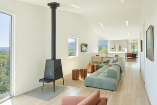 Interior view of a living area at Triple Barn house featuring white walls, wood flooring, pastel coloured seating, a tall, black fireplace, windows, wall art, a light wood unit with items on top and small wooden side tables