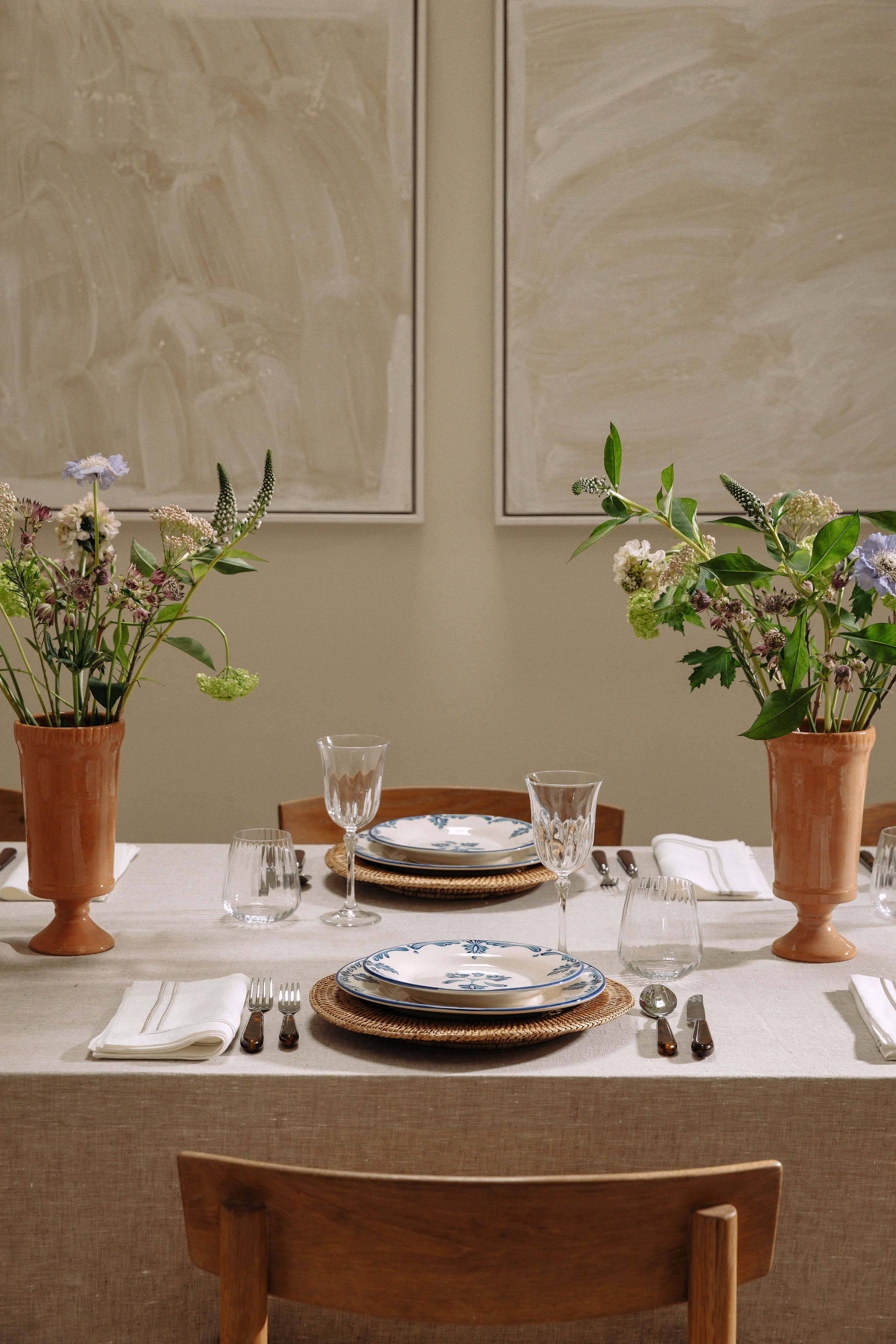 A modern, minimalistic dining table, styled with a beige table cloth and terracotta vases of wildflowers as centre pieces. The place settings consist of vintage style plates and crystal wine glasses.