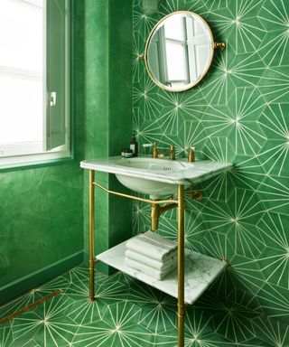 Bright green bathroom with green, Moroccan style floor and wall tiles, textured green painted walls, rounded mirror with glass wall lights ether side, traditional basin with white sinks and lower white shelf, brass metalwork