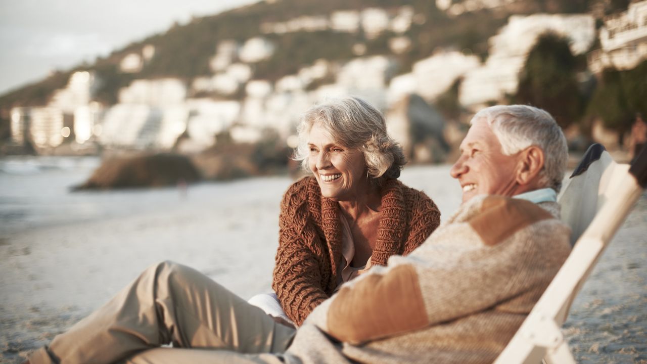 A retired couple smile while relaxing on deck chairs at a beach. 