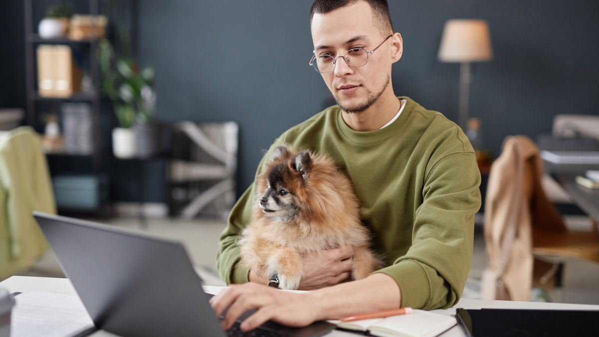 A man sitting at a table while using a laptop and holding a dog