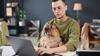 A man sitting at a table while using a laptop and holding a dog