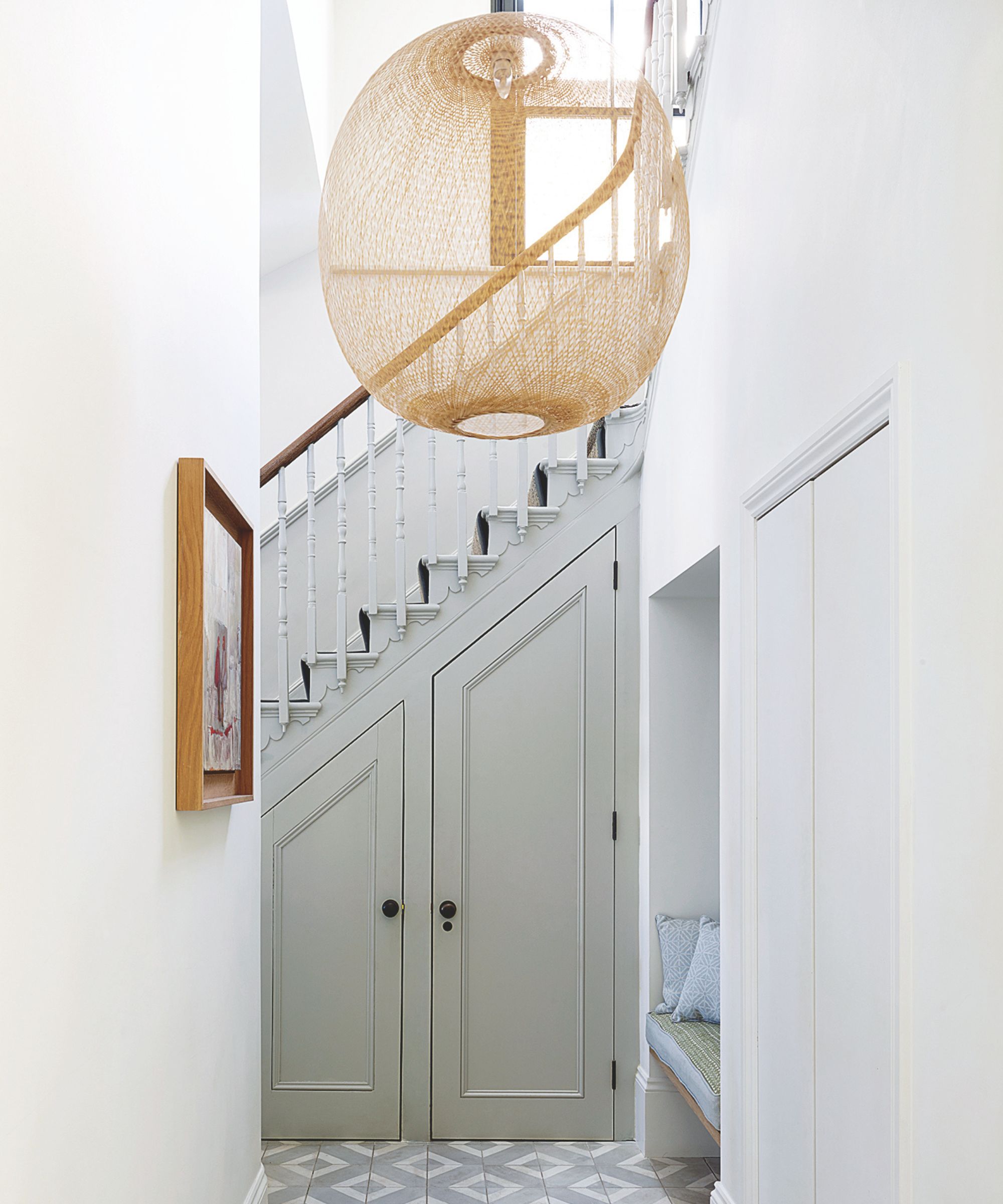 A gray understair cupboard in a bright entryway with white walls. The staircase has white posts with a wooden banister. The floor is gray and white tiled.