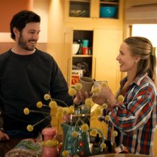 A man and a woman (Adam Brody as Noah and Kristen Bell as Joanne) smile while standing in a kitchen in front of a bouquet of yellow flowers, in 'Nobody Wants This' season 3.