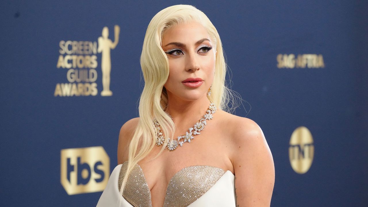 Lady Gaga wearing a white dress with her blonde hair down, standing in front of a navy blue background with gold logos