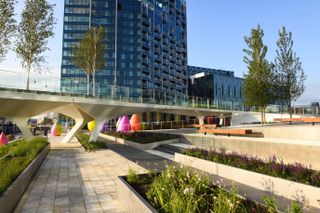Alternative view of The Tide riverside park during the day. The park features a raised walkway, benches, grass, trees, green and purple plants in rectangular planters and colourful waterdrop-shaped structures. There are glass front buildings nearby