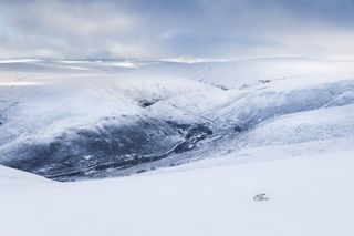 A snow-covered mountainous landscape with a mountain hare captured in the bottom-right corner of the frame