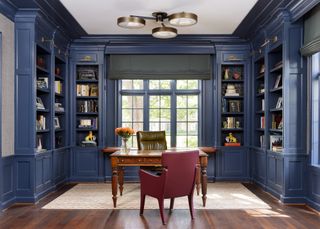 A library painted in Benjamin Moore's Mysterious blue shade with dark wood flooring and a desk and two chairs sitting atop a rug underneath a ceiling light