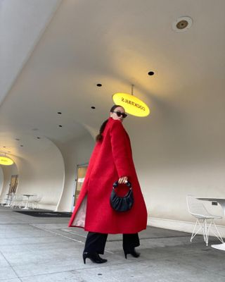 Jasmine Fox-Suliaman wearing red statement coat, black pants, black boots and black phone handle purse.