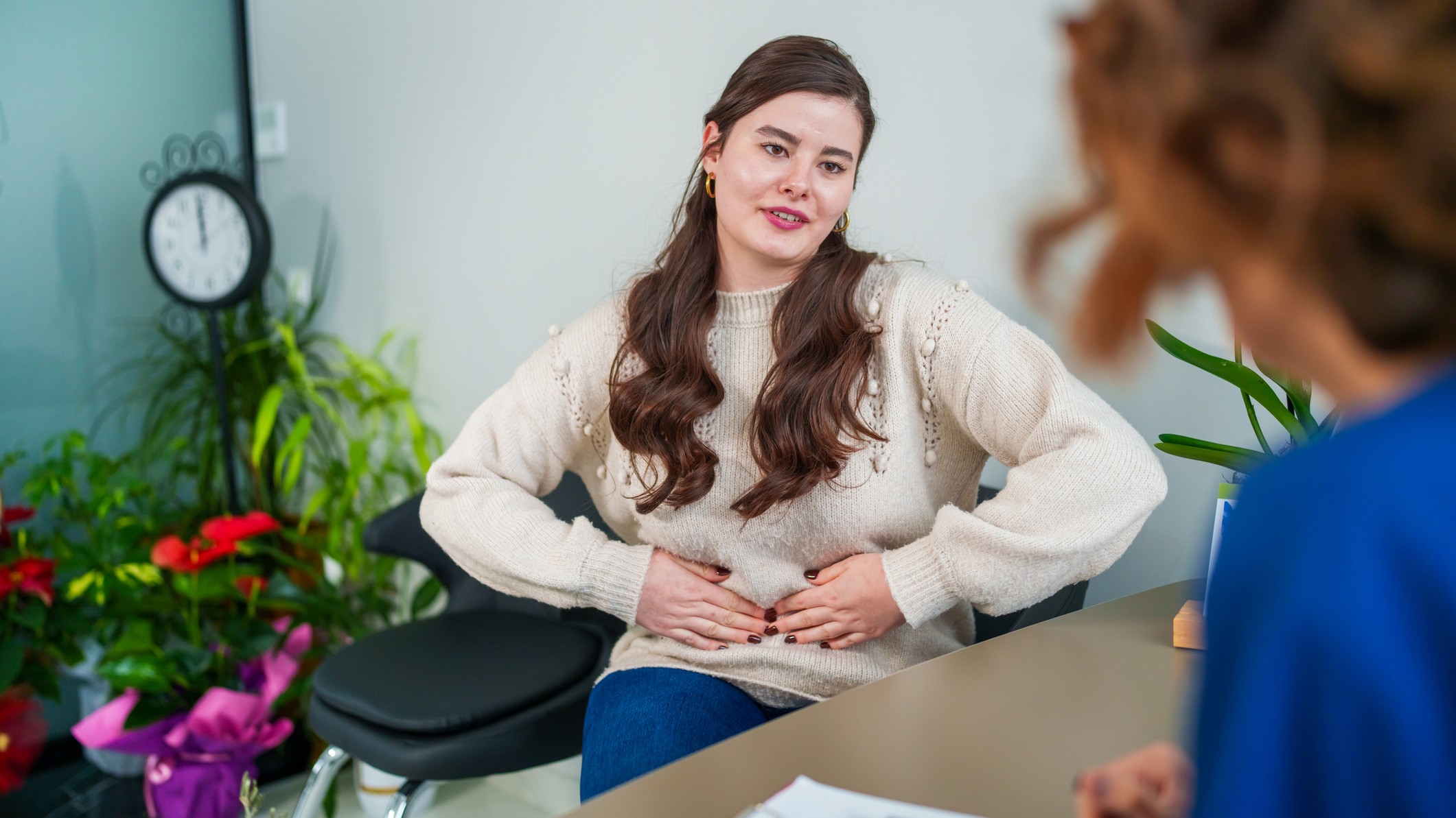 A woman sits behind a desk in an apparently doctor's office and we see her over the doctor's shoulder. She presses her hands on her stomach and is in the middle of the language.