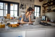 A young, smiling woman works on a traditional IRA on her laptop in her kitchen.