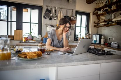 A young, smiling woman works on a traditional IRA on her laptop in her kitchen.