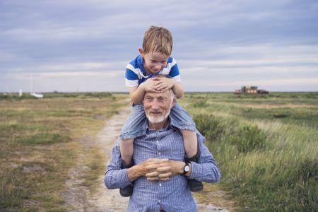 A young boy, hot and sweaty from running around, smiling while being carried by his grandad on an evening walk through a coastal salt marsh.