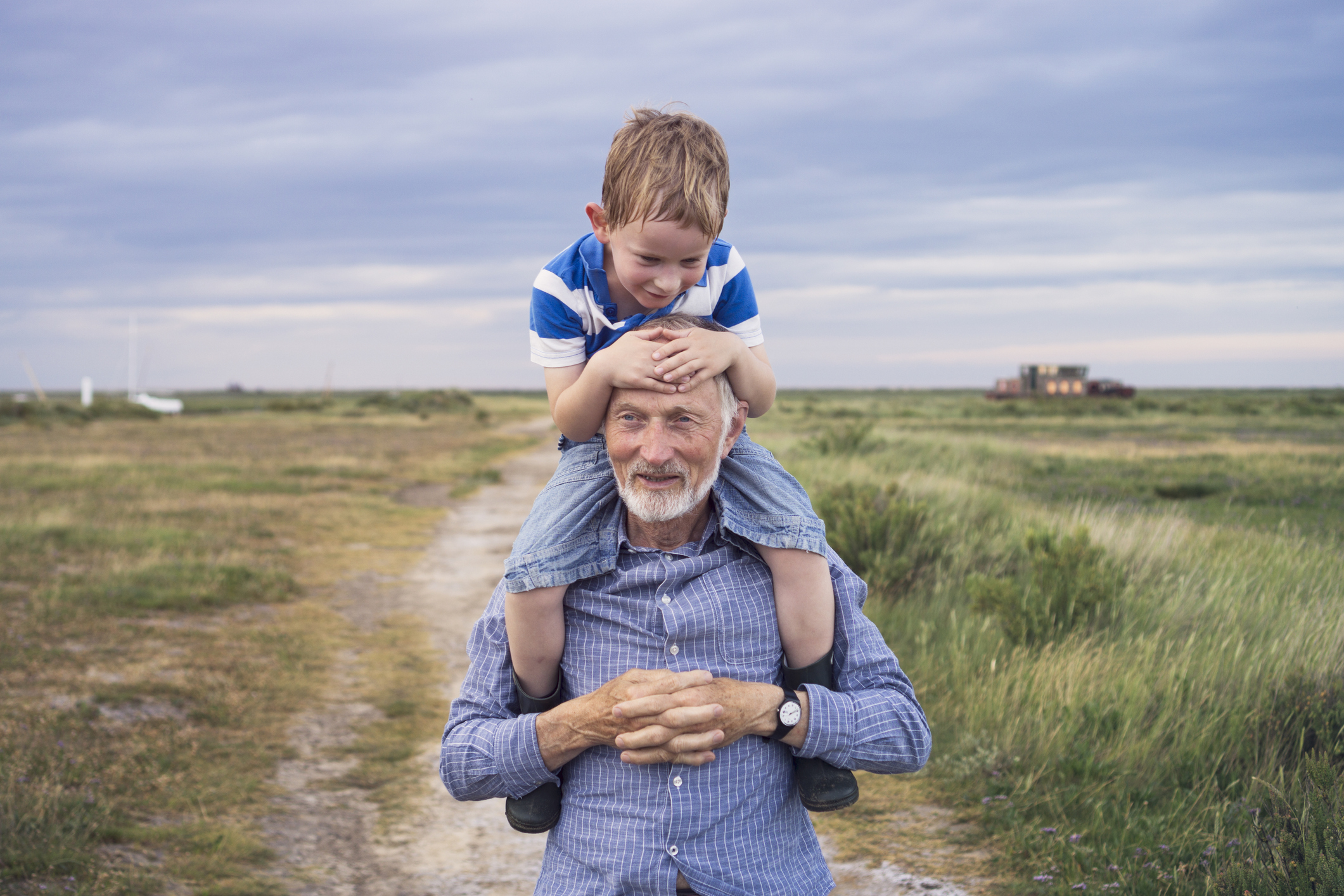 A young boy, hot and sweaty from running around, smiling while being carried by his grandad on an evening walk through a coastal salt marsh.