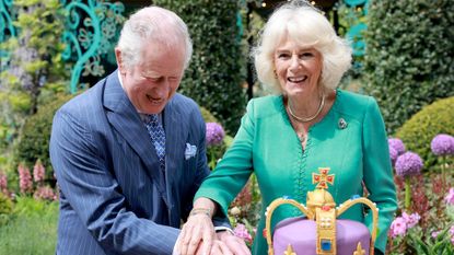King Charles and Queen Camilla laugh while cutting a crown shaped cake