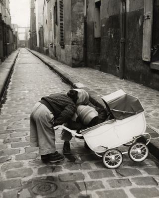 Black and white photograph of a child holding a pram in the middle of a street.
