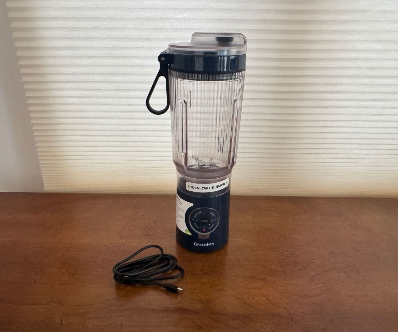 A black based the GreenPan Spin &amp; Go portable blender on a wooden table beside a charging cable.