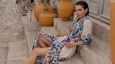 A woman sitting and leaning against stone steps wearing a white and colourful patterned dress from Boden. 