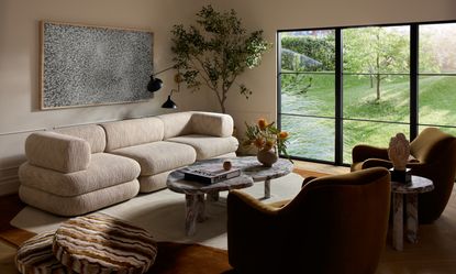 Cream living room with black Crittall-style doors, cream sofa, cream pouffe, wood wavy grey marble coffee table and olive green armchairs