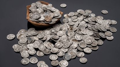 A small wooden bowl filled with silver coins &mdash; and one gold coin. A pile of silver coins is spread in front of it.