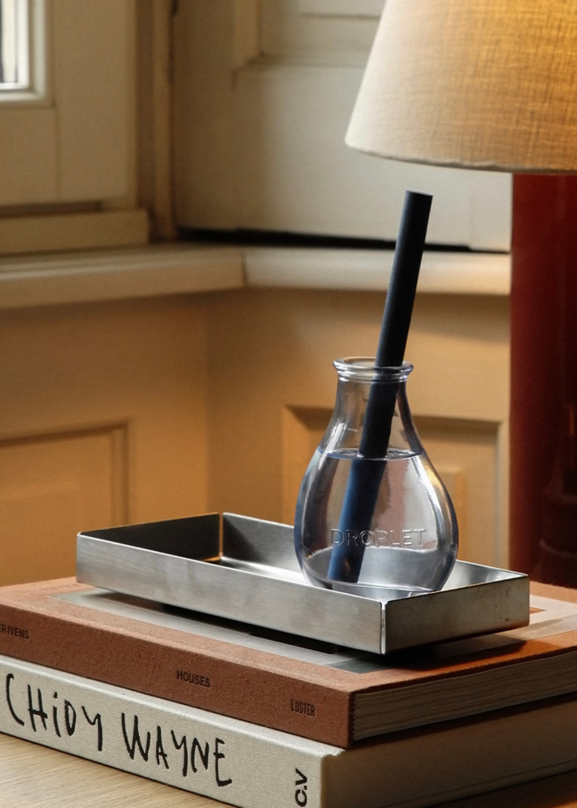 A blue single reed diffuser from Droplet on a steel tray on a stack of coffee table books beside a table lamp