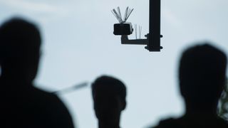 Silhouettes of people walking beneath a facial recognition surveillance camera