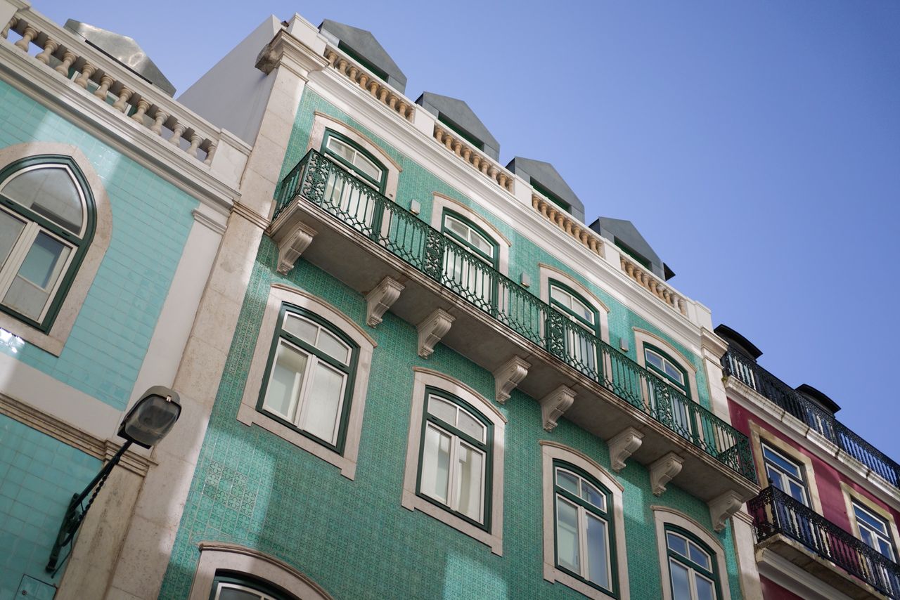 Low-angle view of a turquoise tiled building with ornate balconies and white stone trim against a clear blue sky.