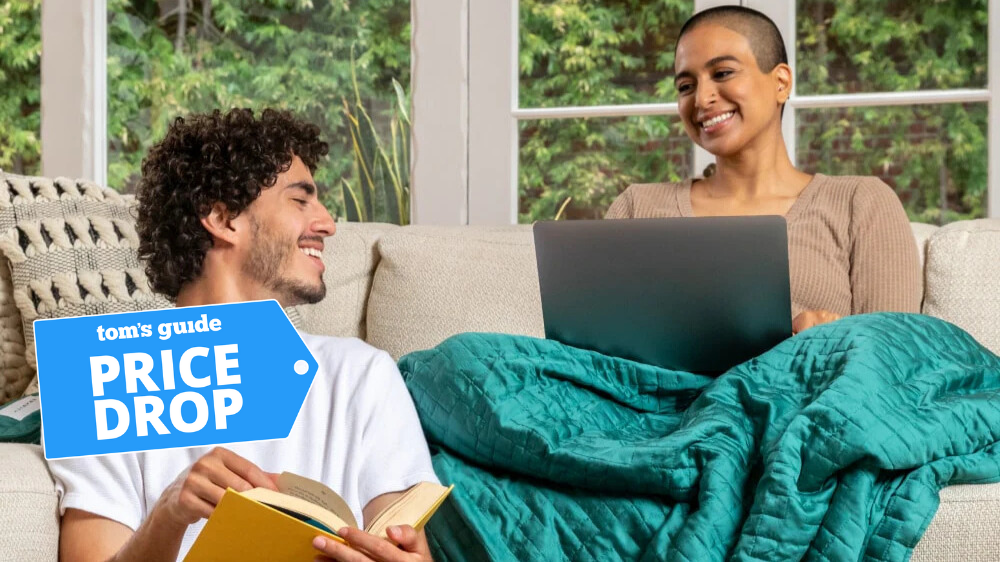 The image shows a man holding a book while looking back at a woman sitting on the couch with a laptop and a green weighted blanket draped on her lap