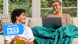 A man holding a book staring at a woman sitting on the couch with a laptop and a green shining blanket draped on her lap