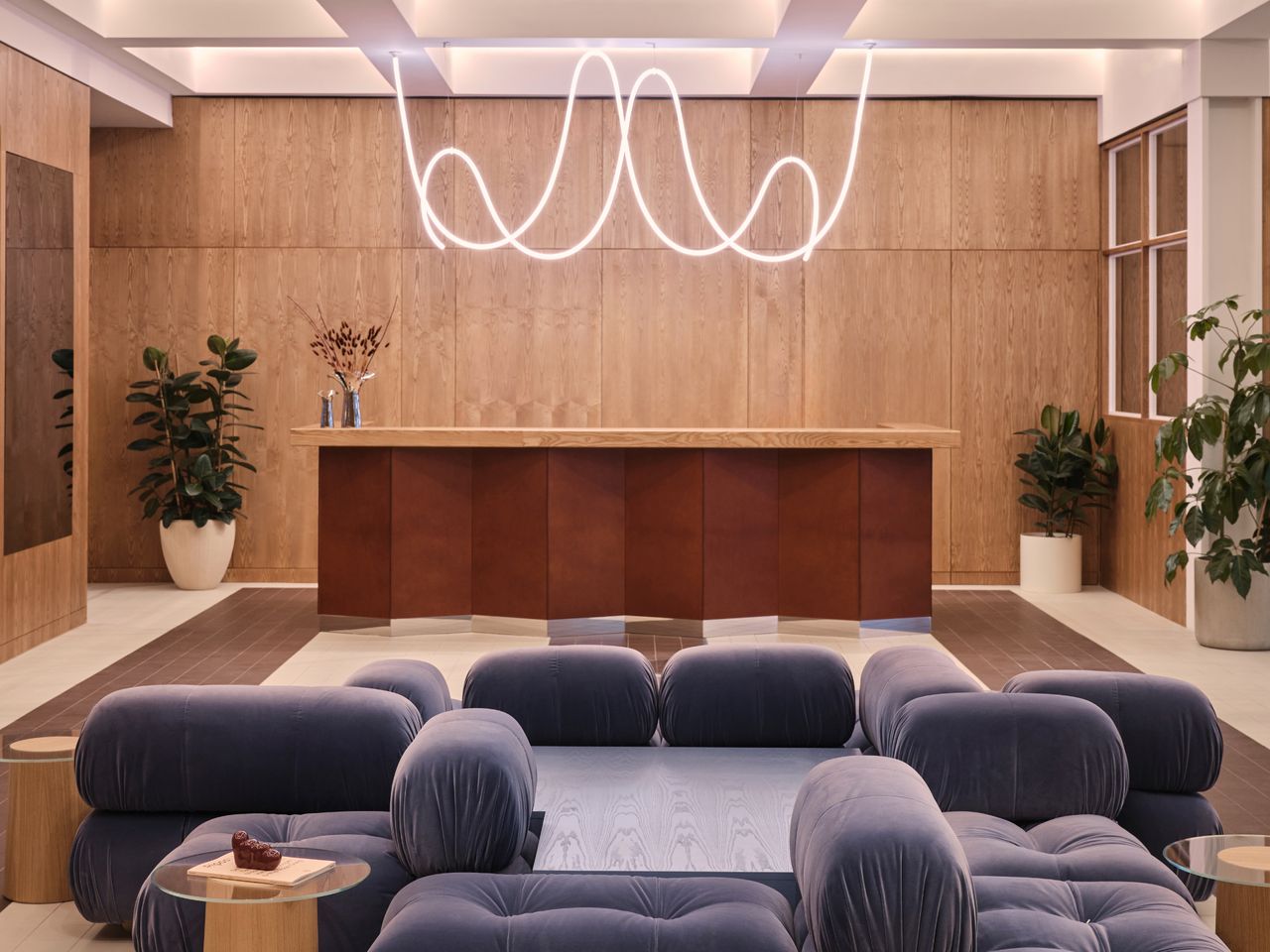 Lobby area featuring grey sofas back-to-back with a table inbetween; a large front reception desk with large in-door plant pots in the room&#039;s corners. A wave squiggle illuminated light hangs from above. 