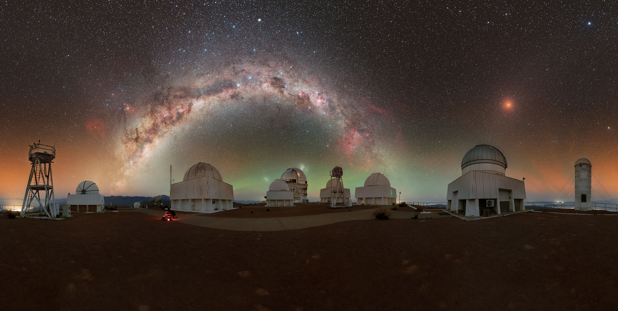 A panoramic view of observatories under a starry sky, featuring the vibrant Milky Way and a reddish celestial body in the background