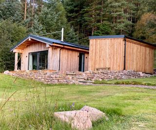 A timber-clad holiday home situated on a gently sloping plot on a Scottish croft, surrounded by woodland. The single-storey building, created by Norscot, has a dual pitched roof and modern patio doors installed in the gable end. A facilities block sits adjacent and is also clad in timber.