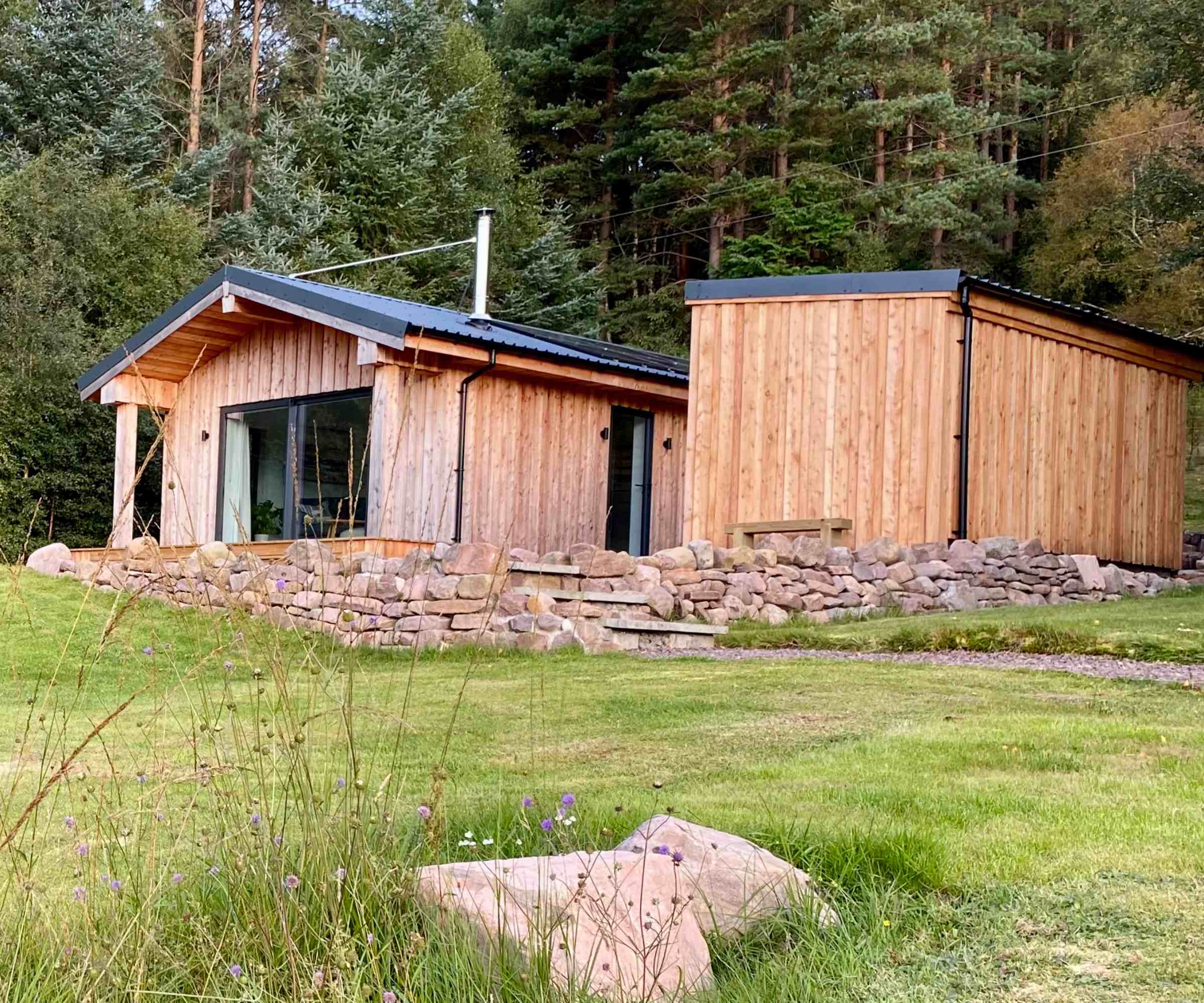 A timber-clad holiday home situated on a gently sloping plot on a Scottish croft, surrounded by woodland. The single-storey building, created by Norscot, has a dual pitched roof and modern patio doors installed in the gable end. A facilities block sits adjacent and is also clad in timber.