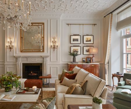Luxurious living room with white panelled walls, artwork and wall lamps, cream sofa and coffee table in fron of fireplace, windows and green seating to the right