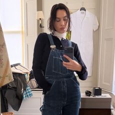 Alexa Chung takes a mirror selfie wearing dark wash dungarees with black rounded-toe boots and white turtleneck jumper layered under a black cardigan.