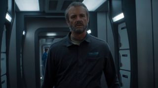 A man with short gray hair and beard wearing a gray jumpsuit is striding down a gray metal corridor. He looks worried.