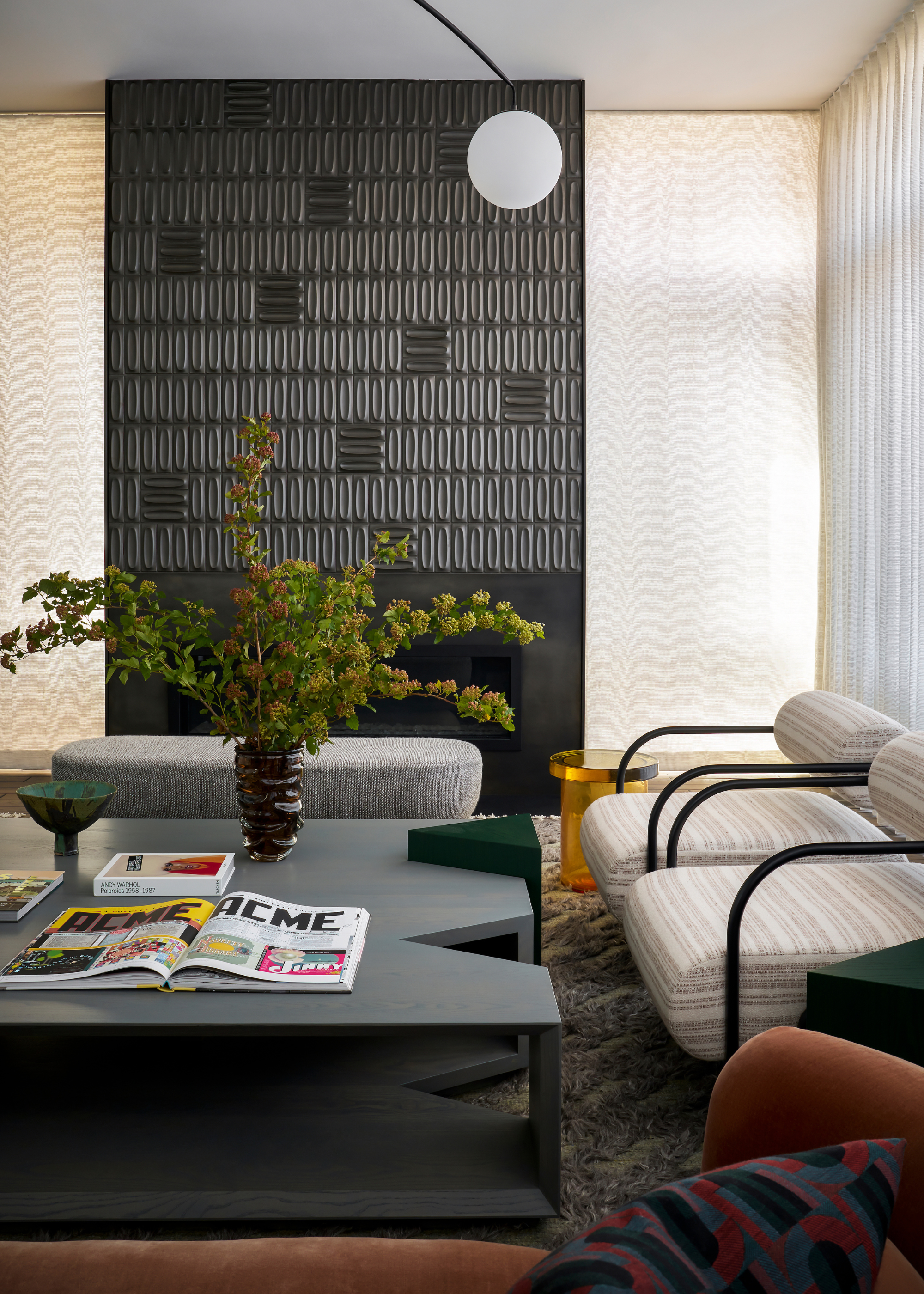 A white living room with a black textured accent wall, a gray bench, a charcoal-colored coffee table with open books, a base of stems, beside a pair of black metal armchairs with striped cream upholstery