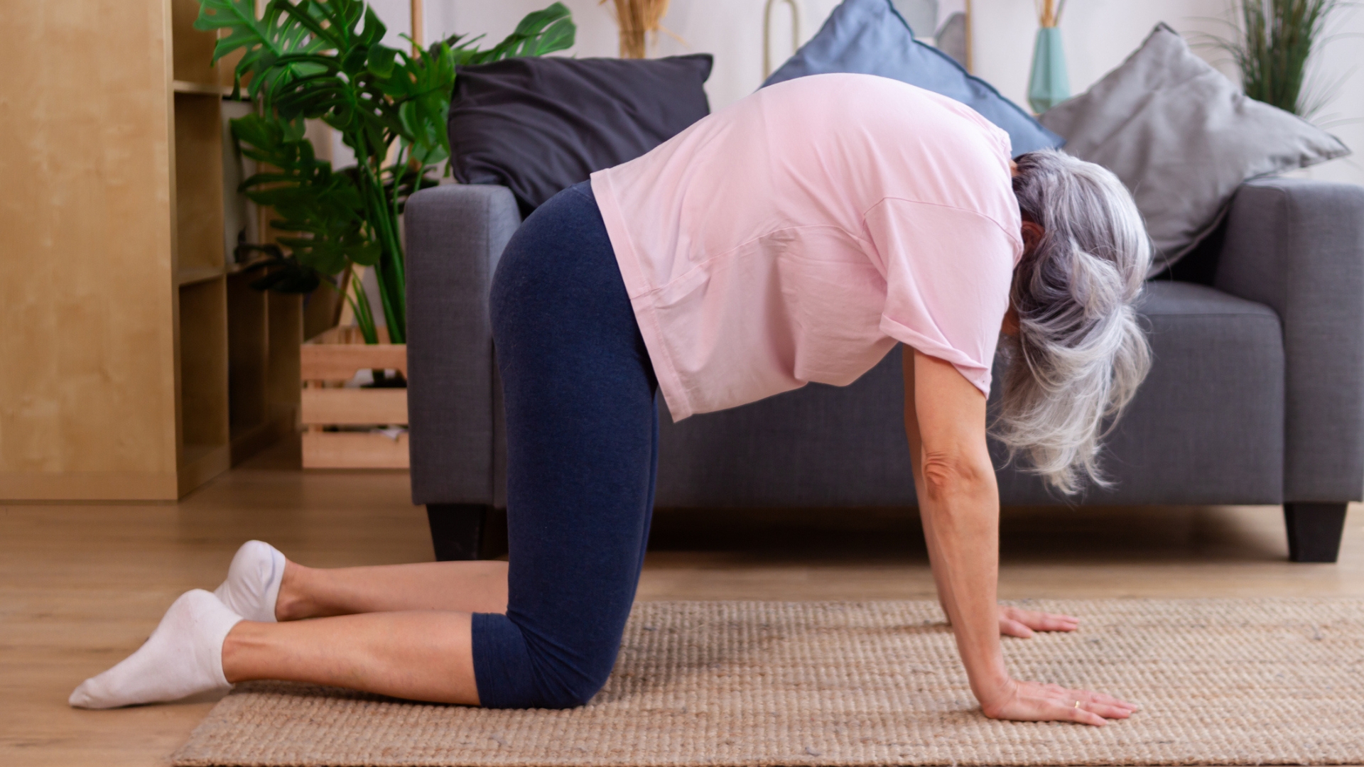 woman sideways to the camera on her hands and knees with spine rounded up and looking down. there's a sofa behind her. she's wearing navy leggings and a pink tshirt.