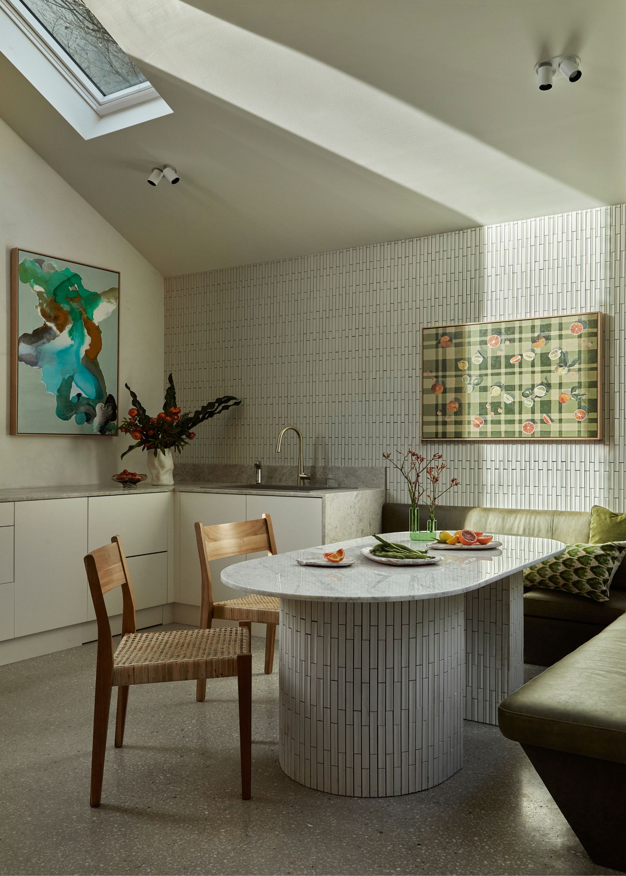 A bright kitchen and dining space with a skylight, a tiled table, wallpapered walls, a framed painting, banquette seating, and rattan chairs