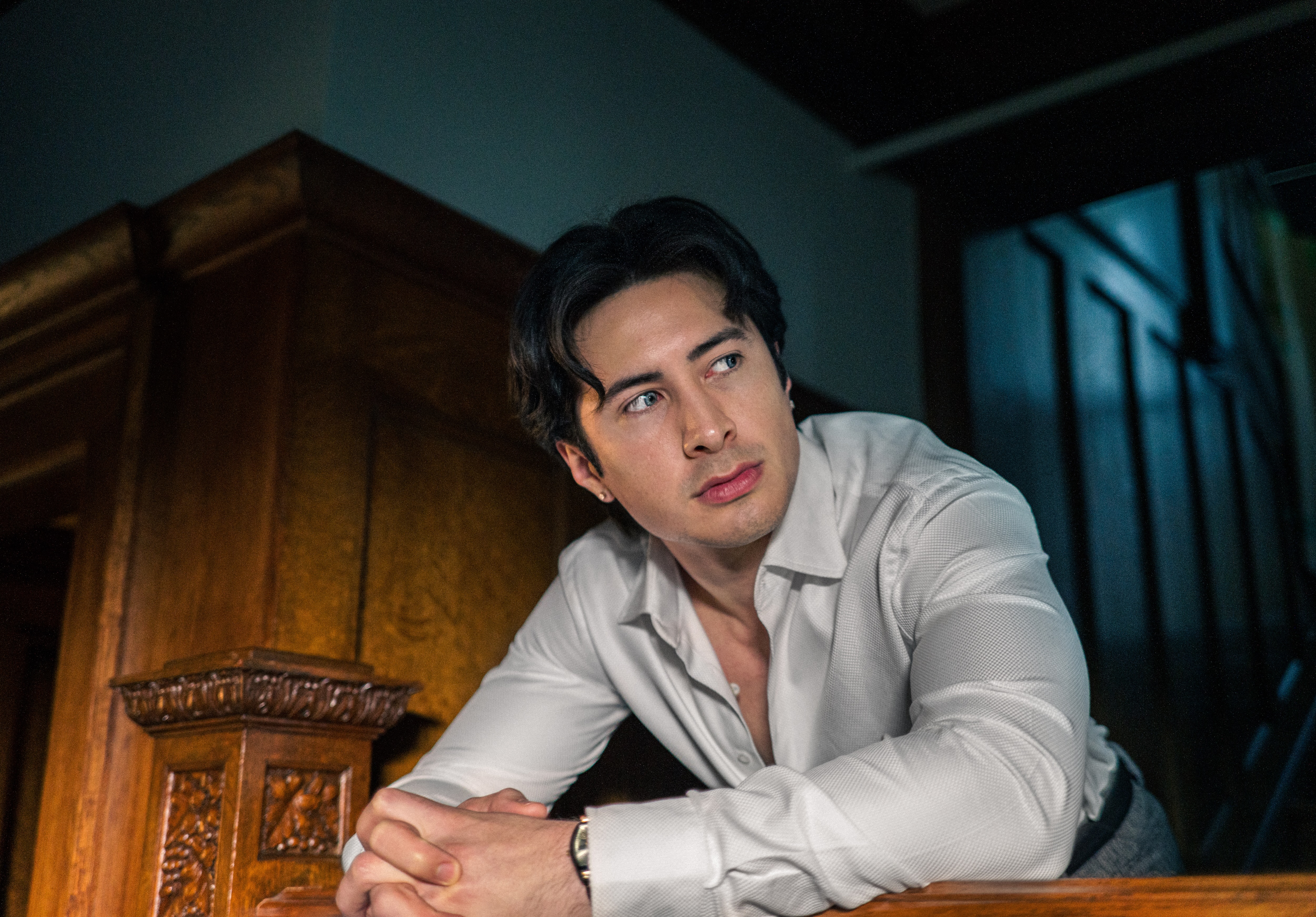 Hudson Williams wears icy blue contact lenses and a white button-down while leaning on wooden furniture in the Crave TV show 'Yaga.'