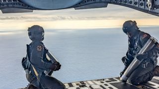 two paratroopers sit at the end of a plane's open cargo hold, looking out over the open ocean